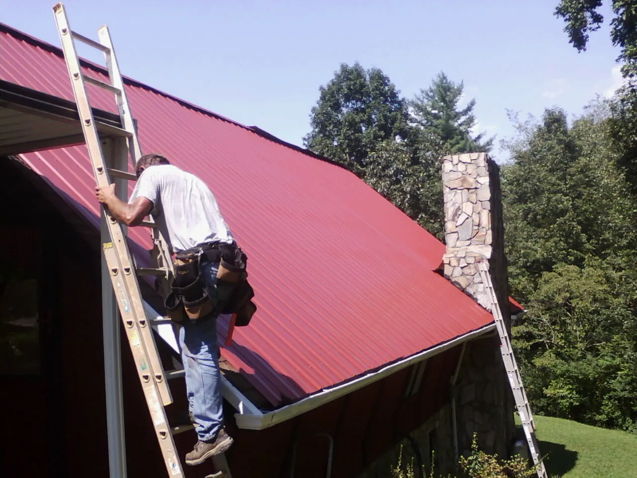 Roofer on a ladder beside a red metal roof and stone chimney — Soffit & Fascia Repair by Roofing Brothers in Hooven